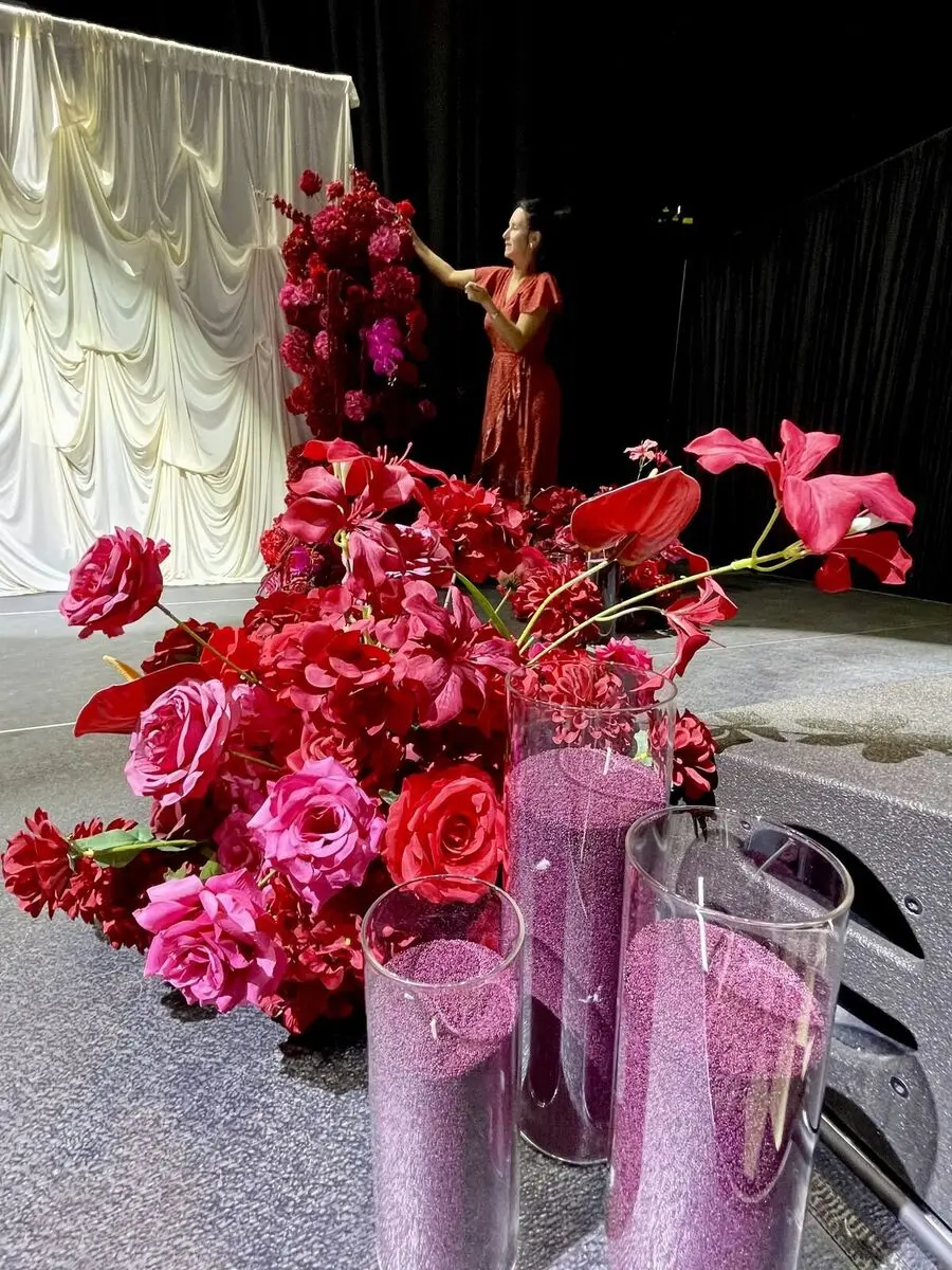 Florist arranging a deep red and pink floral installation on a stage with white draped backdrop.